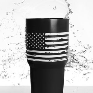 Peut inclure: Un gobelet en acier inoxydable noir avec un motif de drapeau am&eacute;ricain d&eacute;lav&eacute; blanc. Le gobelet est entour&eacute; de gouttelettes d'eau.