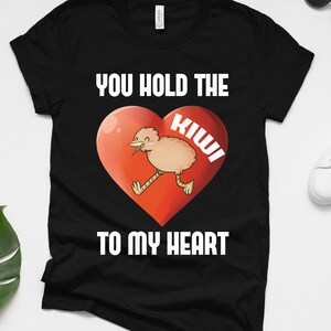 Op de afbeelding: Zwart T-shirt met een rood hart en een cartoon kiwi erin. De tekst "You hold the kiwi to my heart" staat op het shirt gedrukt.