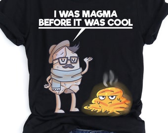 May include: Black t-shirt with a cartoon illustration of a rock wearing a hat and scarf, and a puddle of molten lava. The text "I was magma before it was cool" is printed on the shirt.
