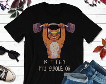 Op de afbeelding: Zwart T-shirt met een cartoon oranje kitten dat gewichten heft. De tekst "Kitten My Swole On" is onder het kitten gedrukt.