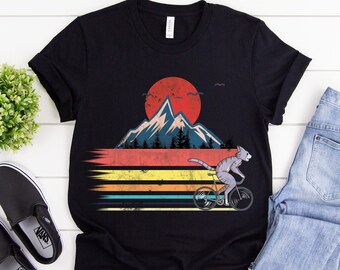 May include: Black t-shirt featuring a retro design of a gray cat riding a bicycle in front of a mountain and sunset. The design includes horizontal stripes in yellow, orange, and blue. The shirt is paired with blue jeans and black slip-on shoes.