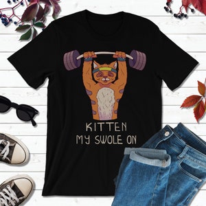 Könnte beinhalten: Schwarzes T-Shirt mit einem Cartoon-orangefarbenen Kätzchen, das Gewichte hebt. Der Text "Kitten My Swole On" ist unter dem Kätzchen aufgedruckt.
