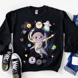 Puede incluir: Sudadera negra con un gráfico de un mono con un casco espacial flotando en el espacio. El mono está rodeado de planetas, estrellas y donas.