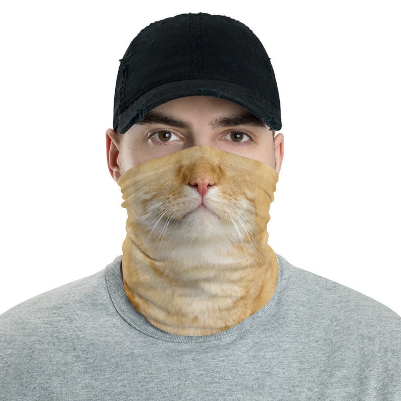 May include: A person wearing a black baseball cap and a face mask with a cat's face printed on it. The cat's face is light orange with a pink nose and white whiskers.