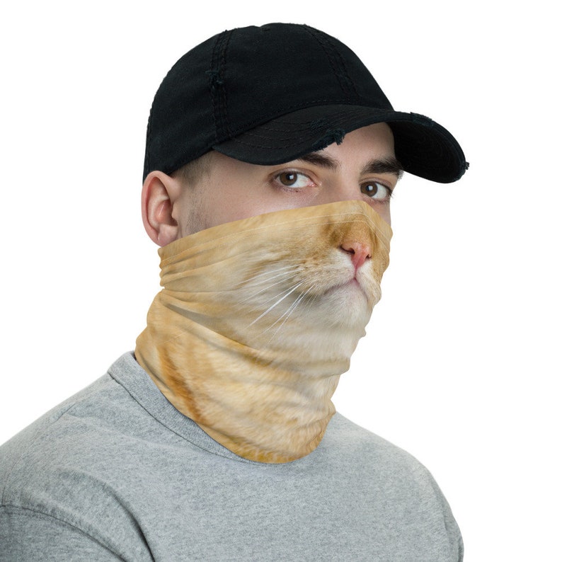 May include: A person wearing a black baseball cap and a face mask with a cat's face printed on it. The cat's face is orange and white.