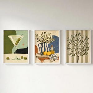 May include: A set of three framed wall art prints. The first shows a martini glass with olives on a table. The second depicts a blue and white striped pitcher with lemons and olive branches. The third features an olive tree on a striped background.