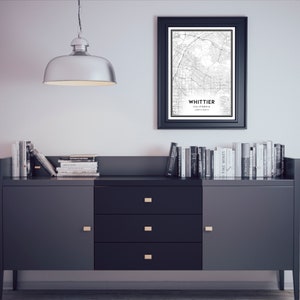 May include: A black and white map print of Whittier, California, framed in a black frame, hanging on a white wall above a dark grey dresser with three drawers. The dresser has gold-colored handles and is topped with stacks of books.