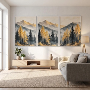 Peut inclure: Un ensemble de trois tableaux muraux représentant un paysage de montagne avec des sommets brumeux et une forêt d'arbres vert foncé et jaune doré. Les trois cadres sont disposés horizontalement au-dessus d'un meuble télé en bois clair.
