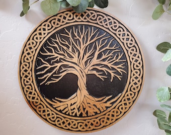 May include: Round wooden wall art featuring a Tree of Life design. The tree is carved in a light wood color against a black background, surrounded by a Celtic knot border. Decorative eucalyptus branches are in the background.
