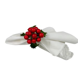 May include: A white fabric napkin folded and secured with a decorative ring featuring a cluster of shiny red berries and dark green holly leaves. The napkin has a smooth texture and a scalloped edge visible on the right side of the folded fabric.
