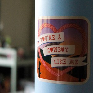 May include: A sticker with a pink heart rope design and the text "You're a cowboy like me" on a light blue background.