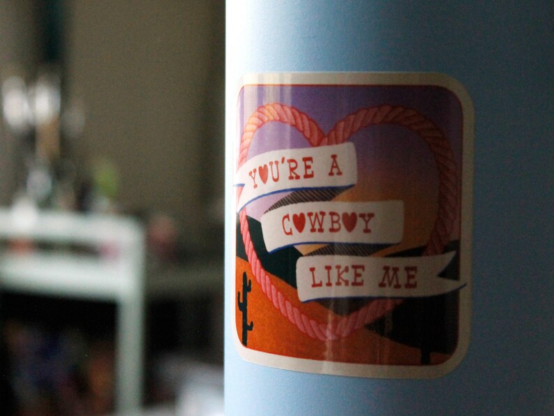 May include: A sticker with a pink heart rope design and the text "You're a cowboy like me" on a light blue background.