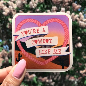 May include: A pink and orange sticker with a rope heart design. The sticker says "You're a cowboy like me" with a heart in place of the "o" in "cowboy".