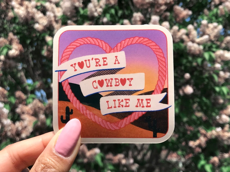 May include: A pink and orange sticker with a rope heart design. The sticker says "You're a cowboy like me" with a heart in place of the "o" in "cowboy".