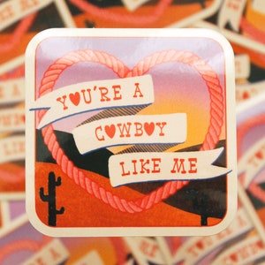May include: A sticker with a pink heart rope design and a sunset background. The sticker says "You're a Cowboy Like Me" with a red heart in each word.
