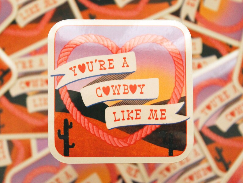 May include: A sticker with a pink heart rope design and a sunset background. The sticker says "You're a Cowboy Like Me" with a red heart in each word.