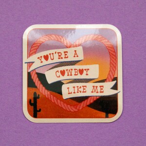 May include: A square sticker with a sunset background and a rope heart. The heart has three banners with the text "YOU'RE A COWBOY LIKE ME" with a heart symbol in each banner.