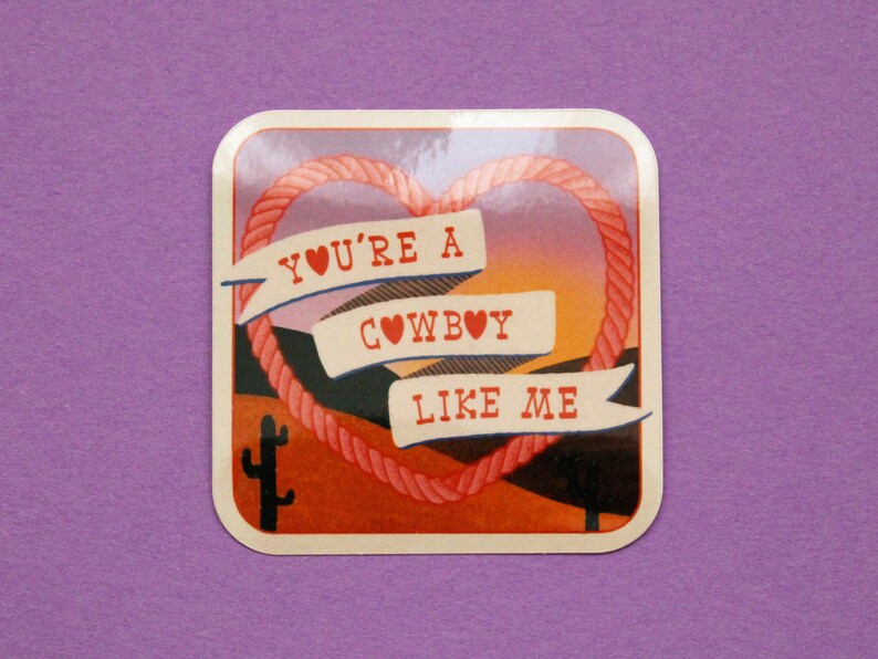 May include: A square sticker with a sunset background and a rope heart. The heart has three banners with the text "YOU'RE A COWBOY LIKE ME" with a heart symbol in each banner.