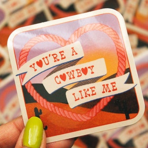 May include: A sticker with a pink heart rope design and a sunset background. The sticker says "You're a Cowboy Like Me" with red hearts in the text.