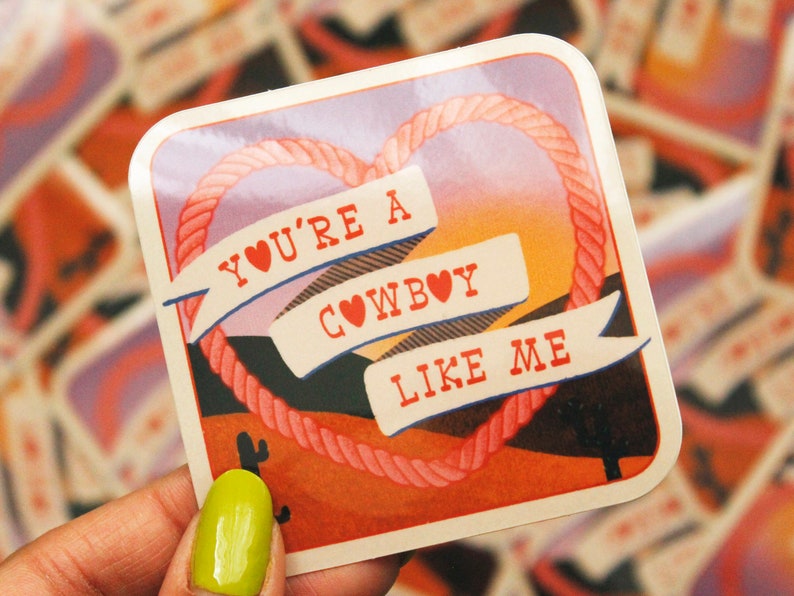 May include: A sticker with a pink heart rope design and a sunset background. The sticker says "You're a Cowboy Like Me" with red hearts in the text.