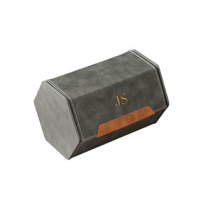 Op de afbeelding: Een grijze zeshoekige doos met een bruine leren klep en de initialen "JS" in goud gegraveerd. De doos is gemaakt van een zacht, su&egrave;de-achtig materiaal.