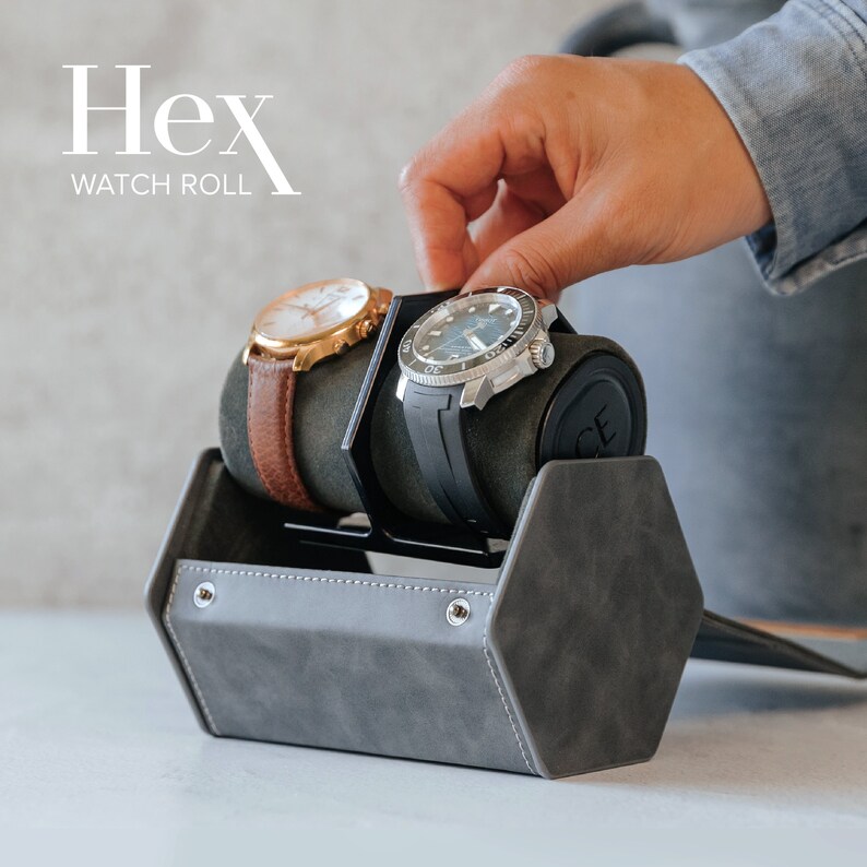 Op de afbeelding: Een grijze zeshoekige horloge-rol met twee horloges erin. De horloge-rol heeft een zwarte leren band en een zilveren gesp. De horloges zijn een zilveren en zwarte horloge met een zwarte band en een gouden horloge met een bruine band.