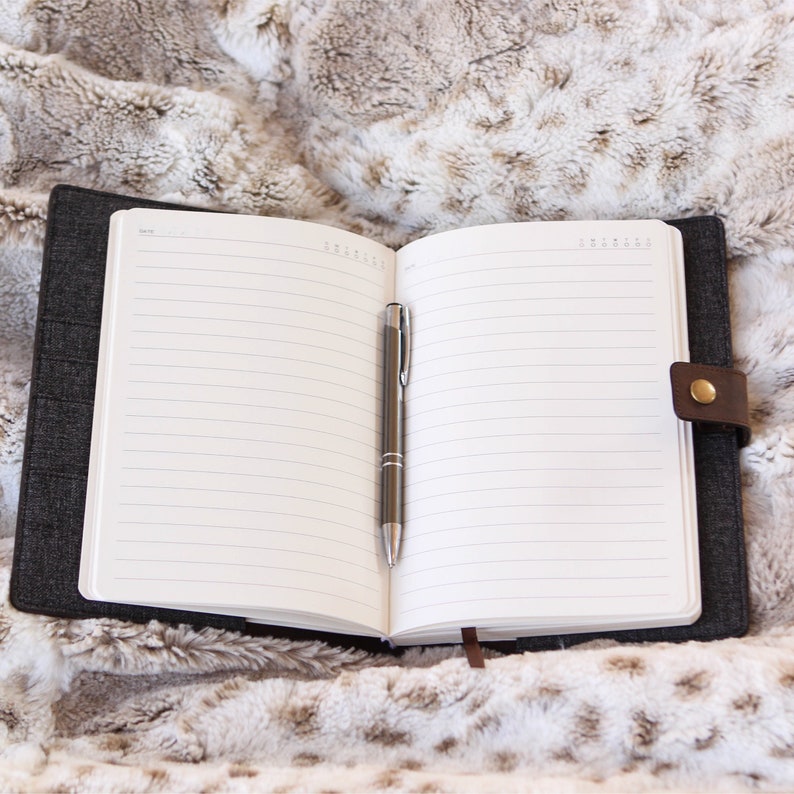 May include: An open black notebook with lined pages and a silver pen lying on a fluffy white and brown faux fur surface.