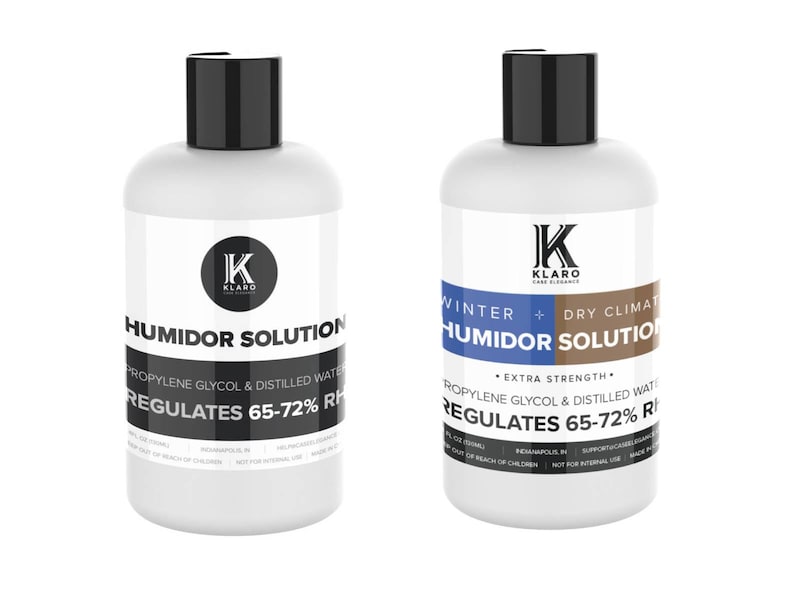 May include: Two white plastic bottles with black caps and labels. The bottles are labeled "Humidor Solution" and contain a solution to regulate humidity levels. The bottle on the left is labeled "Humidor Solution" and the bottle on the right is labeled "Winter + Dry Climate Humidor Solution". Both bottles have the text "KLARO" on the label.