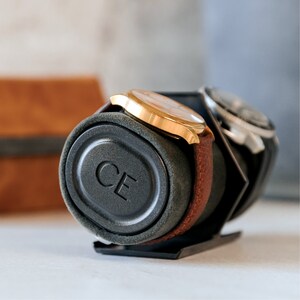 May include: A black watch winder with a gray velvet interior and a gold watch with a brown leather band. The winder has the letters "CE" embossed on the side.