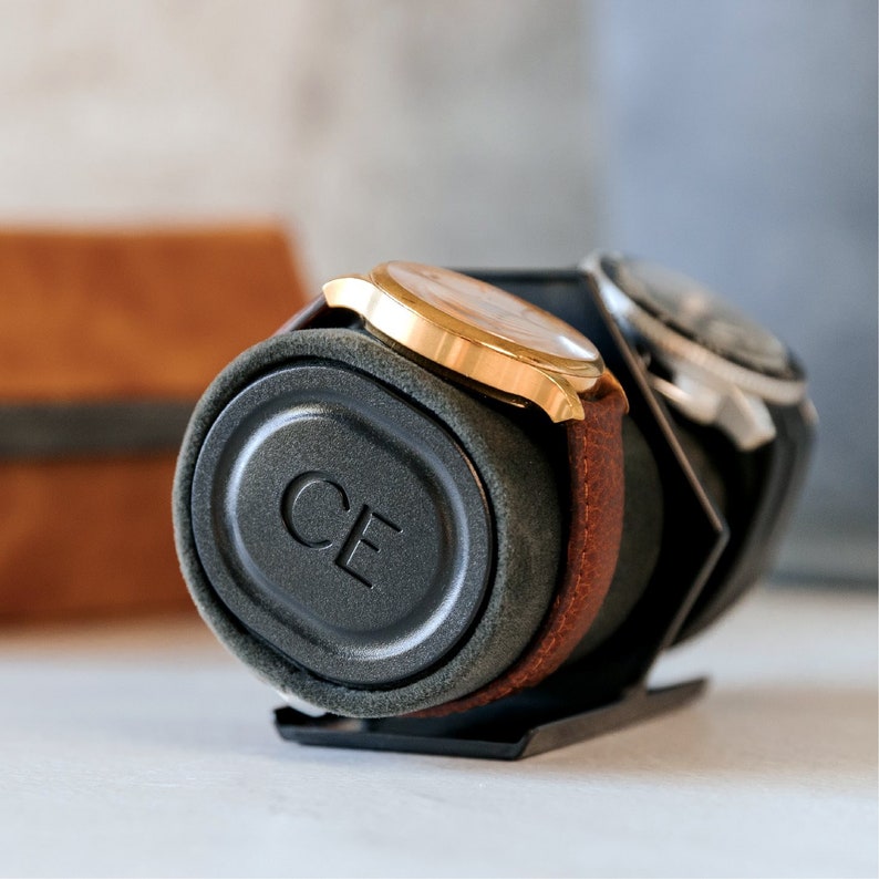 May include: A black watch winder with a gray velvet interior and a gold watch with a brown leather band. The winder has the letters "CE" embossed on the side.