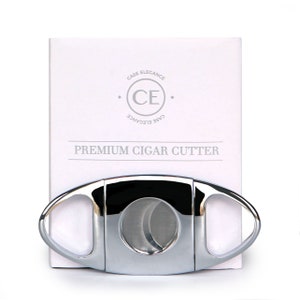 May include: A silver cigar cutter with a curved, oval shape and a sharp blade. The cutter is on a white box with the text "Case Elegance CE Case Elegance Premium Cigar Cutter".