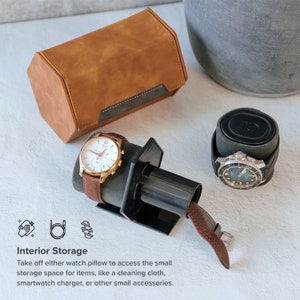 May include: A brown leather watch box with a black interior and a watch winder with a black base and a brown leather strap. The watch winder is holding a silver watch with a white face and black hands. The watch box is sitting on a white surface.