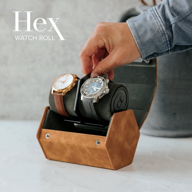 May include: A brown leather watch roll with the text "Hex Watch Roll" on the front. The roll is open and has two watches inside. One watch has a black band and a silver face, and the other watch has a gold band and a white face.
