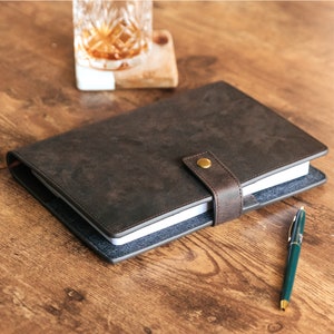 May include: A brown leather-bound journal with a blue denim cover and a brass button closure. The journal is lying open on a wooden table with a pen beside it.