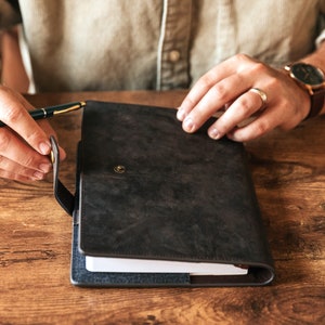 May include: A dark brown leather bound journal with a flap closure and a metal button. The journal is open and the pages are white.