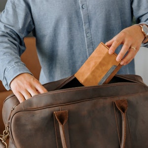 May include: A brown leather duffel bag with a zippered opening. A person is placing a tan leather box with a gray trim into the bag.