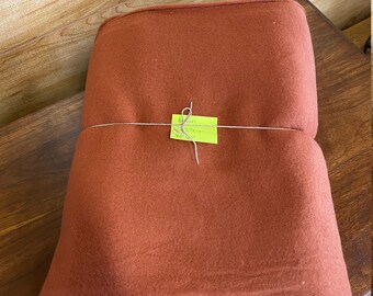 May include: A folded roll of rust-colored fabric, secured with string and a yellow tag. The tag has handwritten text. The fabric is displayed on a dark wooden surface with a lighter wooden background.