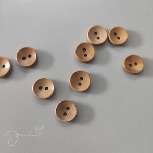 Könnte beinhalten: Zehn Holz-Knöpfe mit jeweils zwei Löchern. Die Knöpfe sind hellbraun und haben eine leicht abgerundete Form.