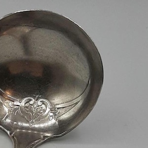 以下が含まれることがあります： ハンドルに装飾的なデザインが施された銀製のサービングスプーン。スプーンは丸いボウルと長く細いハンドルを持っています。