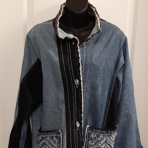 Può includere: Giacca di jeans dal design unico. La giacca presenta un mix di denim blu e pannelli in tessuto nero. Ha due tasche frontali con un motivo decorativo, un colletto nero e una chiusura con bottoni. I bordi della giacca hanno un bordo sfilacciato.