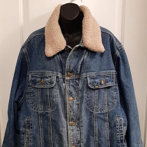 Puede incluir: Una chaqueta vaquera azul con cuello de borreguillo beige. La chaqueta tiene dos bolsillos en el pecho, dos bolsillos laterales y una parte delantera abotonada. La chaqueta está colgada de una percha negra contra una puerta blanca.