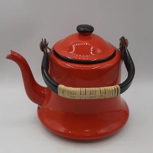 Könnte beinhalten: Eine rote Emaille-Teekanne mit schwarzem Griff und Deckel. Der Griff hat einen geflochtenen Rattangriff. Die Teekanne hat einen Ausguss und einen abgerundeten Körper. Es ist ein Küchenartikel.