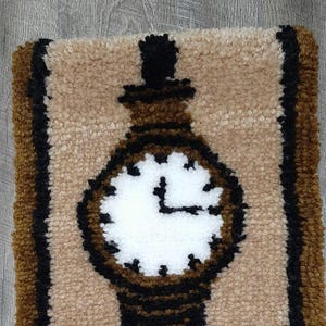 Könnte beinhalten: Ein brauner und schwarzer Teppich mit einem Uhr-Design. Das Zifferblatt der Uhr ist weiß mit schwarzen Zeigern und Zahlen. Der Teppich hat einen Fransenrand.