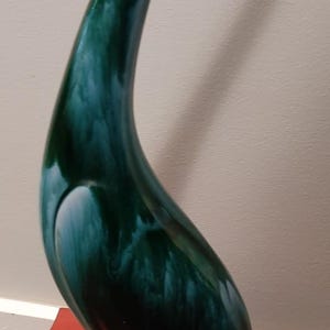 May include: A tall, stylised bird figurine featuring a long, curved neck and a smooth, glossy finish. The body displays a marbled pattern of deep forest green and teal blue hues, transitioning into a solid, rounded black base at the bottom.