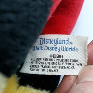 May include: Close-up of a vintage Disney plush toy. The tag reads "Disneyland Walt Disney World" and includes the Disney copyright. The toy is made of black, red, and yellow fabric.