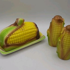 May include: A ceramic butter dish and salt and pepper shakers set, all shaped like corn on the cob. The butter dish is yellow with green and brown detailing, on a green rectangular base. The shakers are corn-shaped, with brown and green accents.