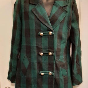 May include: A green and black plaid coat with a double-breasted design. The coat features gold-coloured buttons and two front pockets. The coat is displayed on a black mannequin.