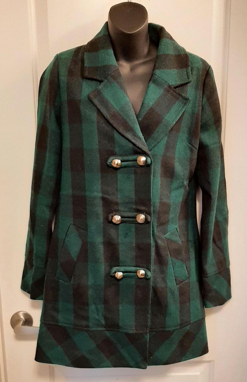 May include: A green and black plaid coat with a double-breasted design. The coat features gold-coloured buttons and two front pockets. The coat is displayed on a black mannequin.