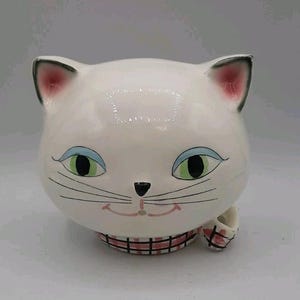 Puede incluir: Un recipiente de cerámica blanca con forma de gato con una cara sonriente, ojos verdes y el interior de las orejas rosado. El recipiente tiene un collar a cuadros rosa y negro y un zapato pequeño a juego. La cara del gato está detallada con bigotes negros y una nariz negra.