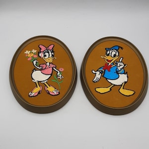 May include: Two embroidered oval framed pictures of Donald Duck and Daisy Duck. Donald Duck is wearing a blue sailor shirt and a sailor hat. Daisy Duck is wearing a pink dress and a pink bow. Both pictures have a brown background.